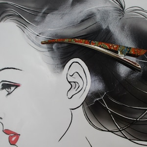 May include: A silver-toned metal hair clip featuring a curved, elongated design with a vibrant, multi-coloured pattern in shades of orange, green, and blue. The clip has a smooth, polished finish on the underside and a decorative top surface.