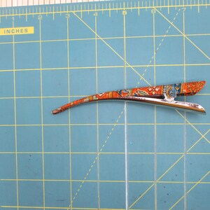 May include: A silver-toned metal hair clip with a curved, elongated shape, featuring an orange, blue, and white patterned fabric overlay. The clip is positioned on a green cutting mat with yellow grid lines, measuring approximately 11.5 cm long.