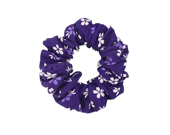 May include: A circular fabric hair scrunchie in a deep purple color featuring a repeating pattern of white and light purple cherry blossom flowers. The accessory has a gathered, ruffled texture throughout the entire circumference of the band.
