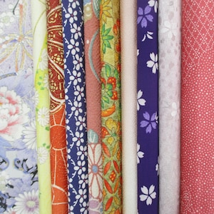 May include: A collection of rolled fabric bolts with various floral and geometric patterns. The fabrics feature colors like blue, red, yellow, and pink. Some bolts have white floral designs, while others have intricate patterns with gold accents.