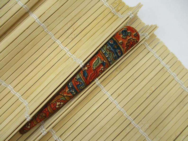 May include: A long, tapered hair stick featuring a fabric-covered surface with a vibrant, multi-coloured floral and geometric pattern in shades of orange, blue, and green, resting diagonally across a light-coloured bamboo mat surface.