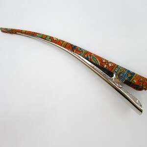 May include: A long, curved hair clip featuring a metallic silver base and a fabric-covered top section. The fabric displays a vibrant, intricate pattern in shades of orange, blue, green, and white, reminiscent of a traditional woven rug design.