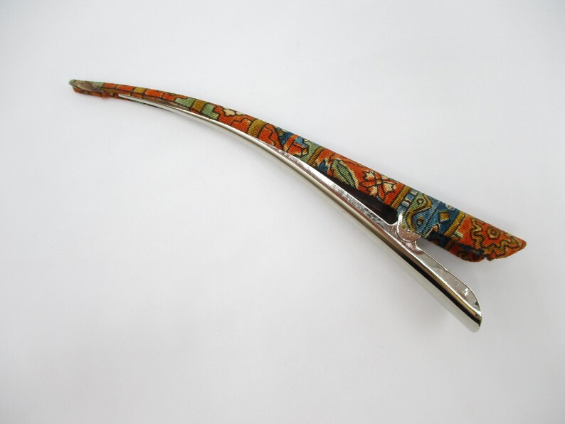 May include: A long, curved hair clip featuring a metallic silver base and a fabric-covered top section. The fabric displays a vibrant, intricate pattern in shades of orange, blue, green, and white, reminiscent of a traditional woven rug design.