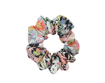 May include: A fabric hair scrunchie featuring a black background with a vibrant, multi-colored floral and leaf pattern. The design includes various shades of pink, orange, green, and white, creating a dense, repeating botanical print throughout the accessory.
