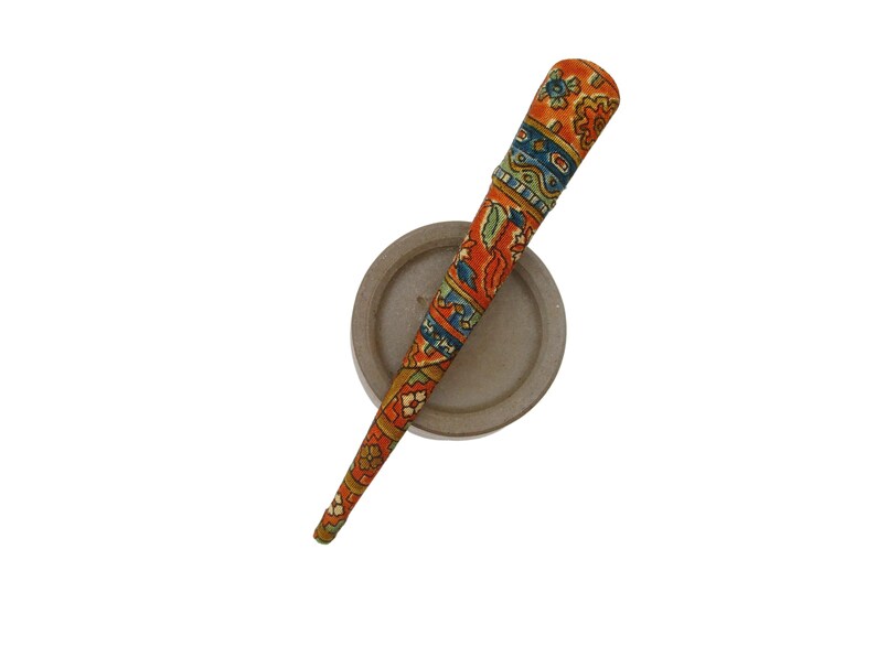 May include: A tapered hair stick featuring a vibrant orange base colour with intricate, multi-coloured floral and geometric patterns in shades of blue, green, and yellow, resting diagonally across a plain, circular, light grey disc-shaped base.