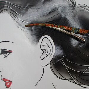 May include: A silver-toned metal hair clip featuring a curved, elongated design with a vibrant, multi-coloured pattern in shades of orange, green, and blue. The clip has a smooth, polished finish on the underside and a decorative top surface.