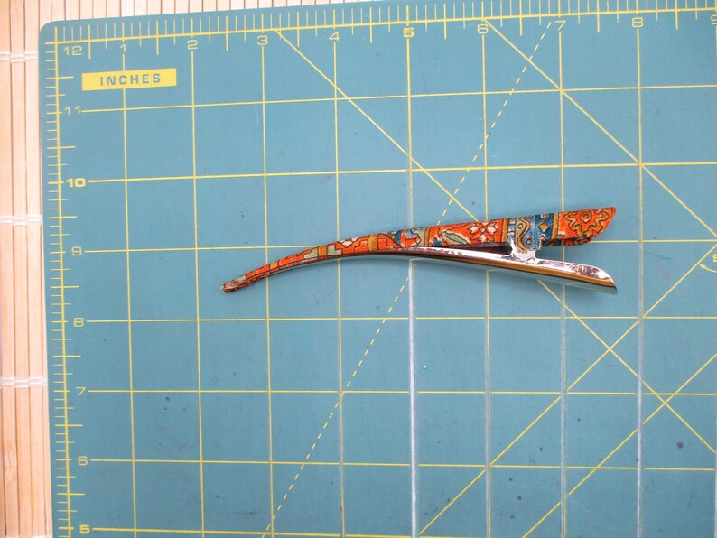 May include: A silver-toned metal hair clip with a curved, elongated shape, featuring an orange, blue, and white patterned fabric overlay. The clip is positioned on a green cutting mat with yellow grid lines, measuring approximately 11.5 cm long.