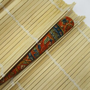 May include: A long, tapered hair stick featuring a fabric-covered surface with a vibrant, multi-coloured floral and geometric pattern in shades of orange, blue, and green, resting diagonally across a light-coloured bamboo mat surface.
