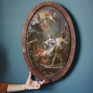 May include: An oval-shaped framed painting depicting a religious scene with a woman in a white robe surrounded by figures in a fiery setting. The painting is set against a blue wall.