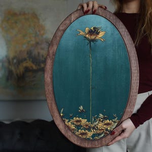May include: A wooden oval frame with a teal blue background and a gold painted flower with a bee on it. The flower is surrounded by smaller gold flowers.