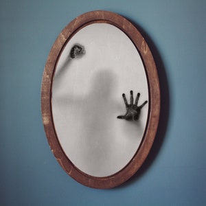 May include: Oval mirror with a dark wooden frame. The mirror's surface reflects a shadowy figure and two black hands reaching out. The background is a solid blue wall.