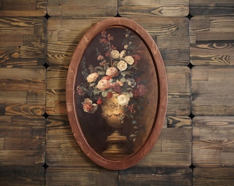 Puede incluir: Una pintura ovalada enmarcada que representa un jarrón lleno de flores rosas, blancas y rojas. El jarrón es dorado y tiene un diseño detallado. La pintura está colocada sobre un fondo de madera marrón oscuro.
