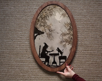 Peut inclure: Une illustration encadrée d'une sorcière et d'un chat assis à une table dans un décor de forêt. L'image est en noir et blanc avec un cadre en bois brun.
