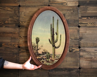 May include: A framed oval print of a desert landscape with a tall cactus in the center. The cactus is green with red flowers and is surrounded by smaller cacti and desert plants. The background is a blue sky with clouds.