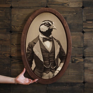 Puede incluir: Un retrato ovalado enmarcado de estilo vintage de un pájaro con un traje y una corbata de lazo. El pájaro tiene plumas blancas y negras y un pico marrón. El retrato está sobre un fondo de madera oscura.