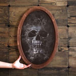 May include: A black and white skull with smoke coming out of its head, framed in a dark wood oval frame.