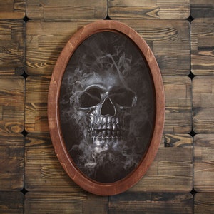 May include: A silver skull with a smoky background is framed in a dark wood oval frame.