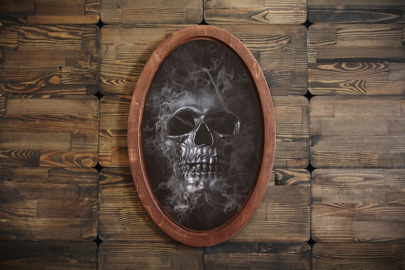 May include: A silver skull with a smoky background is framed in a dark wood oval frame.