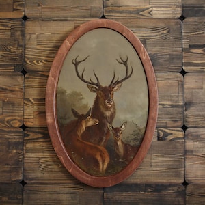 Op de afbeelding: Een ovaalvormig houten frame met een schilderij van een hert en twee kalfjes in een bosrijke omgeving. Het hert heeft een groot gewei en kijkt recht naar de kijker. De kalfjes zijn kleiner en kijken naar het hert.