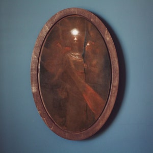 Op de afbeelding: Een ovaalvormig ingelijst schilderij dat drie figuren in een zwak verlichte kamer toont. De figuren staan in een groep, met een heldere lichtbron die het midden van het schilderij verlicht. Het schilderij is ingelijst in een donkerhouten lijst.