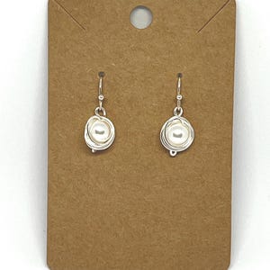 May include: A pair of silver-toned wire-wrapped dangle earrings featuring a single round white pearl in the center of each. The earrings are attached to a rectangular brown cardboard display card with a hole at the top for hanging.