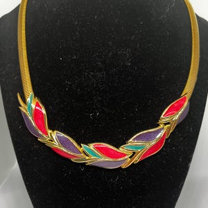 May include: A gold-toned necklace with a flexible chain. The necklace features a series of colorful leaf-shaped pendants in red, purple, and teal, outlined in gold. The necklace is displayed on a black surface.