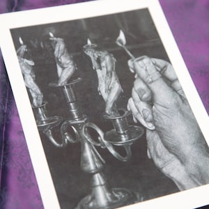 May include: Monochrome print of a candelabra with lit candles. A hand holds a lit match near one of the candles. The image is on a white background, set against a purple patterned fabric.