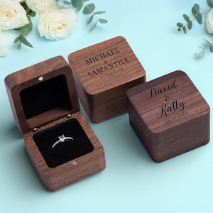 May include: Two personalized wooden ring boxes. The first box is open and shows a black velvet interior with a silver ring. The box is engraved with "Michael & Samantha 10.08.2024". The second box is closed and engraved with "David & Katy 06.21.2024".