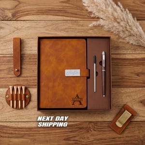May include: A brown leather journal with the name "Austin" engraved on the cover, presented in a gift box with a pen, pen refill, and a money clip. Also included are a leather golf tee holder and a leather strap. The text "NEXT DAY SHIPPING" is visible.