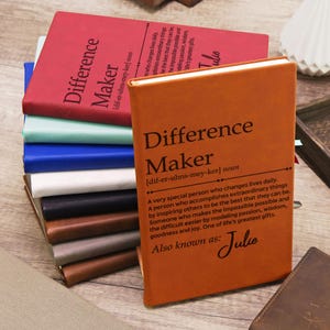May include: A stack of leather-bound journals in various colors, including red, orange, blue, and black. The front journal is orange and features the words "Difference Maker" and a definition. The journals are stacked on top of each other.