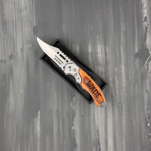 May include: A folding knife with a silver blade and wooden handle, presented in a black box. The blade is engraved with "Groomsman 02-22-2025". The handle is engraved with the name "WADE".
