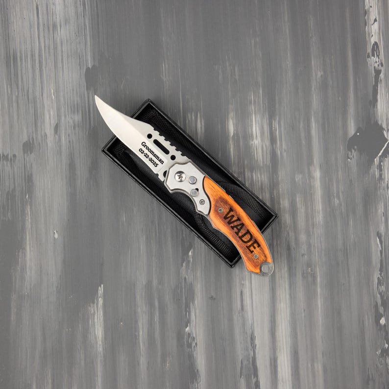 May include: A folding knife with a silver blade and wooden handle, presented in a black box. The blade is engraved with "Groomsman 02-22-2025". The handle is engraved with the name "WADE".