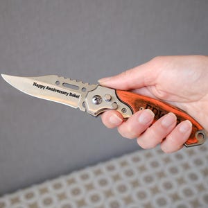 May include: A silver and orange folding knife with the text "Happy Anniversary Babe!" engraved on the blade. The knife has a wooden handle and is held in a hand.