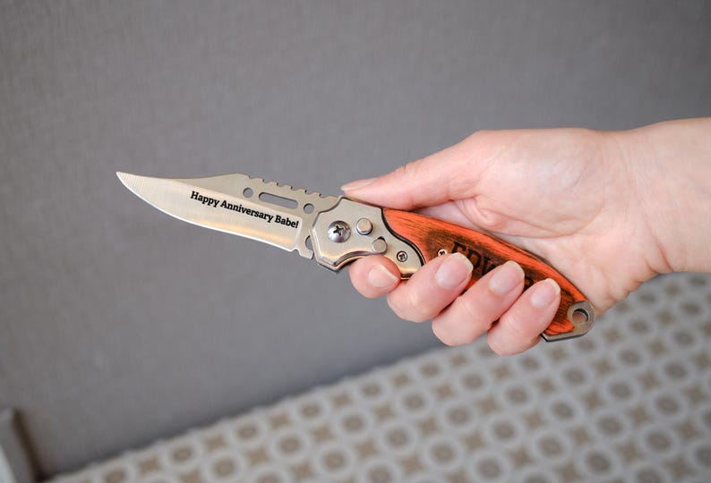 May include: A silver and orange folding knife with the text "Happy Anniversary Babe!" engraved on the blade. The knife has a wooden handle and is held in a hand.
