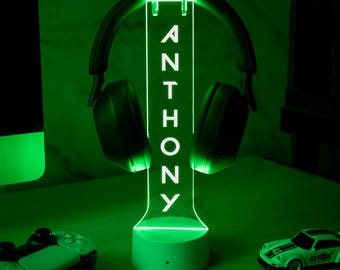 May include: A green LED lamp with the name "ANTHONY" vertically displayed. Black headphones are suspended from the lamp. A green gaming controller and a toy car are on the table. The lamp and objects are illuminated with a green glow.