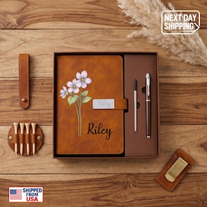May include: A tan leather-like notebook featuring a floral illustration of three light purple flowers and the name Riley printed in black. The set includes a matching pen, a pen refill, a leather golf tee holder with four wooden tees, and a money clip.