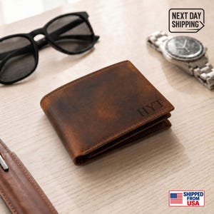 May include: A brown leather bifold wallet featuring the embossed letters HYT on the front corner. The wallet is positioned alongside black sunglasses, a silver-tone analog wristwatch, and a brown leather item with a visible metal edge.