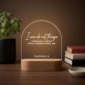 May include: An arched acrylic light display with a wooden base featuring the white text I can do all things THROUGH CHRIST WHO STRENGTHENS ME and PHILIPPIANS 4:13. The clear panel is illuminated with a warm glow, highlighting the engraved lettering.