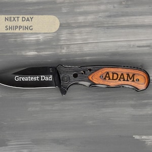 May include: A black folding knife with a wooden handle engraved with the name "ADAM." The blade is etched with the words "Greatest Dad." The knife has a black blade and a wooden handle.