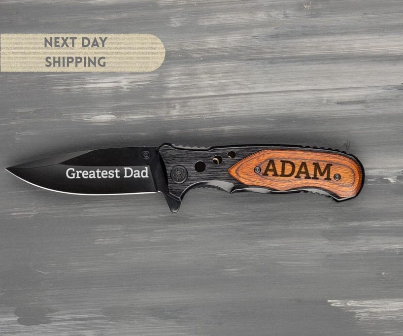 May include: A black folding knife with a wooden handle engraved with the name "ADAM." The blade is etched with the words "Greatest Dad." The knife has a black blade and a wooden handle.