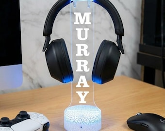 May include: A black headset rests on a clear acrylic stand with the name "MURRAY" illuminated in white. The stand has a blue glowing base and top. A white game controller and black mouse are on the wooden surface.