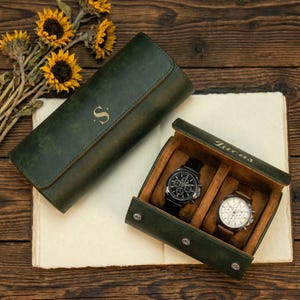 May include: A dark green leather watch roll with the letter S embossed on the exterior and an open watch box containing two wristwatches, one with a black dial and one with a white dial, featuring the name Lucas embossed on the interior lid.