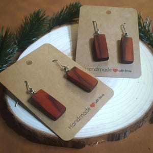 Könnte beinhalten: Rechteckige Holzohrringe mit silberfarbenen Haken, präsentiert auf braunen Karten. Die Ohrringe sind rotbraun mit sichtbarer Holzmaserung. Auf den Karten steht "Handmade with love". Die Ohrringe werden auf einer Holzscheibe präsentiert.