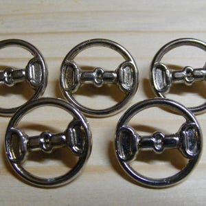 May include: Five silver-colored metal buttons with a circular design and a central, horse-bit-like detail. The buttons are arranged on a light-colored wooden surface. These buttons could be used for clothing or craft projects.