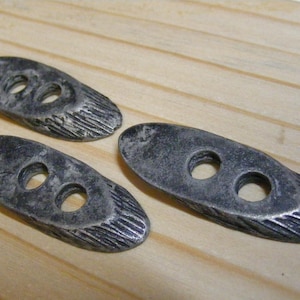 May include: Three oval-shaped, dark gray metal buttons with two holes each. The ends of the buttons have a textured, carved design. The buttons are displayed on a light-colored wooden surface.