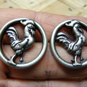 May include: Two silver-toned metal buttons with a rooster design. Each button is round and features a rooster in a circular frame.