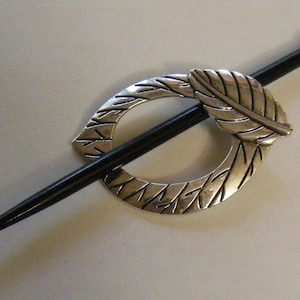 May include: A silver-toned hair pin with a leaf design and a black stick. The pin features a circular design with a leaf accent. The stick is black and has a rounded end. This hair accessory is suitable for various hairstyles.