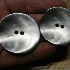 May include: Two large, round, silver-toned buttons with two holes each. The buttons have a slightly textured surface and a vintage appearance. Ideal for sewing projects or adding a unique touch to garments.