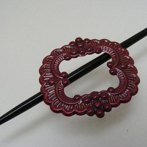 May include: A dark red metal hair stick with a decorative, ornate design. The stick is black and has a rounded end.