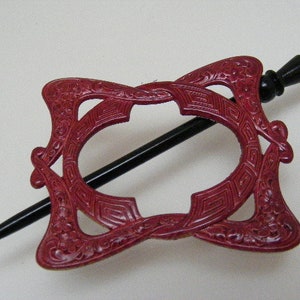 May include: A red plastic hair stick with an intricate design. The stick is shaped like an oval with a black wooden handle.